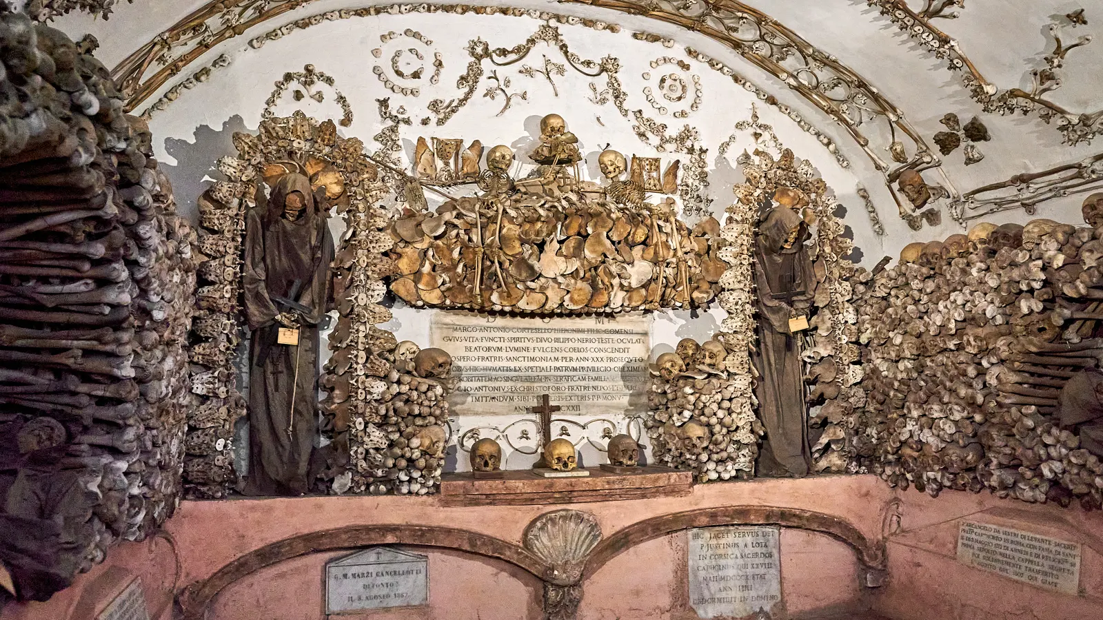 Robed friars and walls of skulls and pelvic bones frame a Latin memorial plaque inside the Capuchin Crypt in Rome