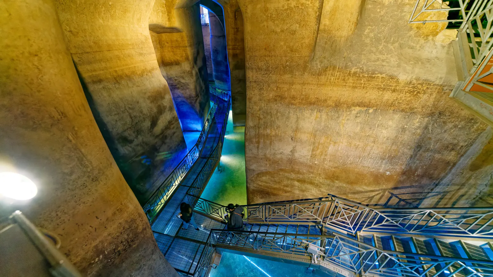 The Palombaro Lungo cistern beneath Matera lit in blue, with a steel walkway crossing the water